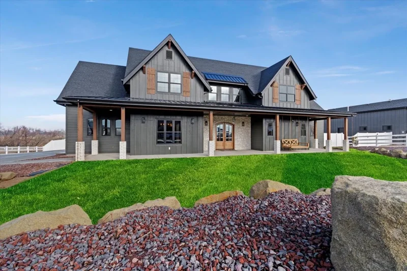A modern two-story house with a dark exterior, wooden accents, and a front porch, surrounded by a green lawn and rocks, under a blue sky.