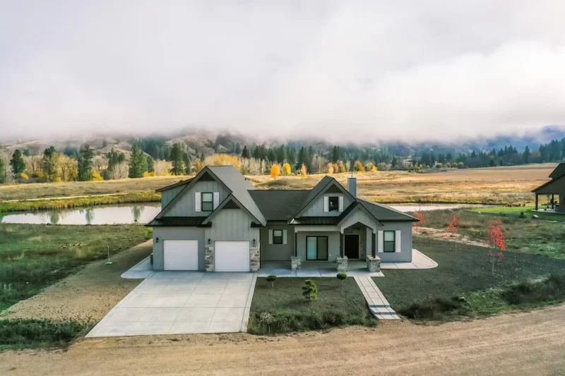 Front exterior of the Gale by Bachman Custom Homes. Light gray exterior with stone, you can see a pond directly behind the home and pine trees and mountains in the distance