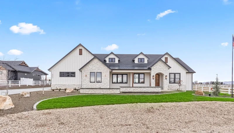 Front view of a modern house with white siding, stone accents, and a dark roof, surrounded by a well-kept lawn and landscaped yard, with a clear blue sky in the background. Located in Nampa's Weatherby Estates Community
