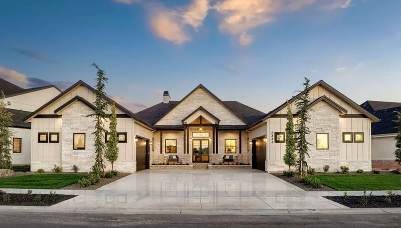 Front Exterior of The Kimberly. Contemporary single-story house with a symmetrical front facade, stone and board siding, three garage doors, and a driveway at dusk with a clear sky.