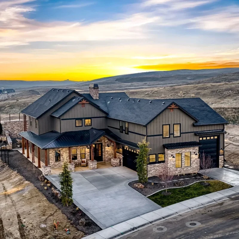 Aerial view of The Bowers in Valnova. Modern house with stone accents, black roof, and a large driveway at sunset with Eagle foothills in the background.
